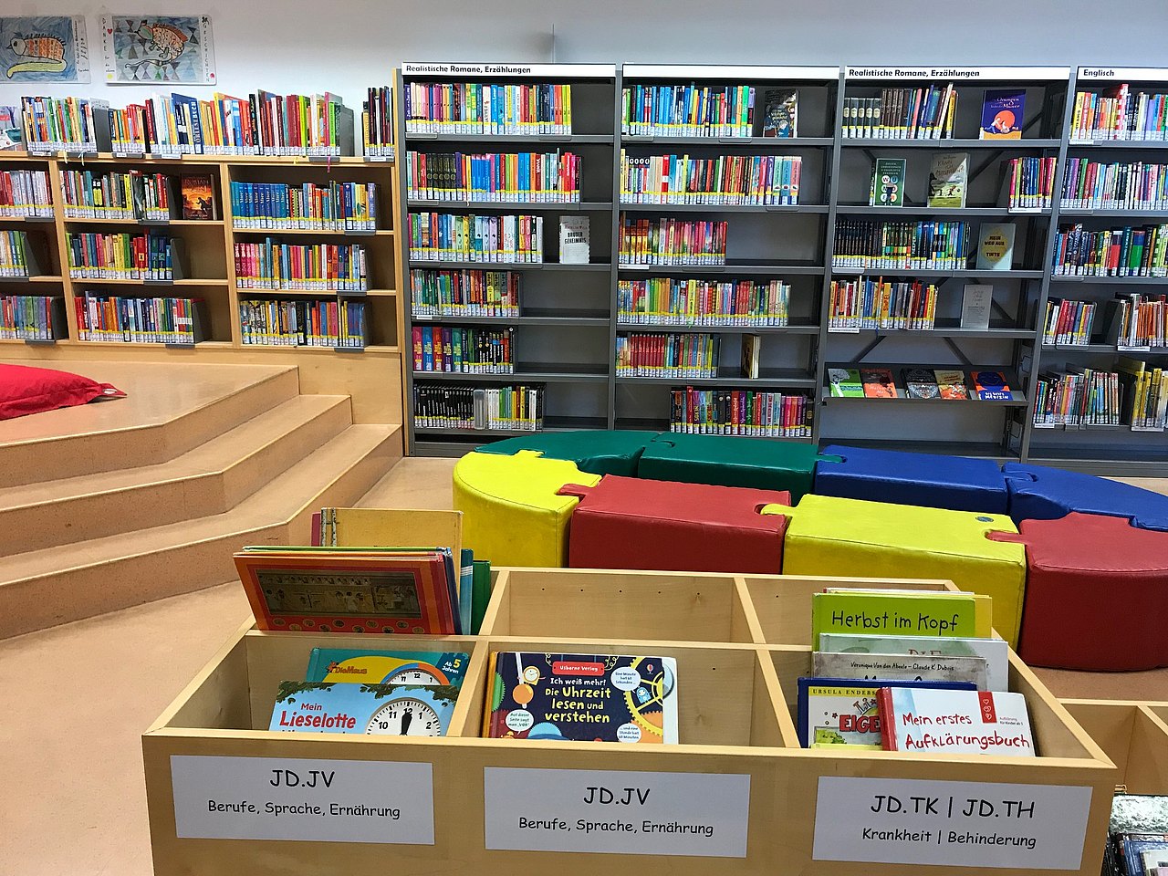 Gegenüber eine Bücherwand, links eine Holzstiege, in der Mitte vom Raum bunte Puzzle-Sitzsteine die ein Oval bilden, davor Bücherkisten.
