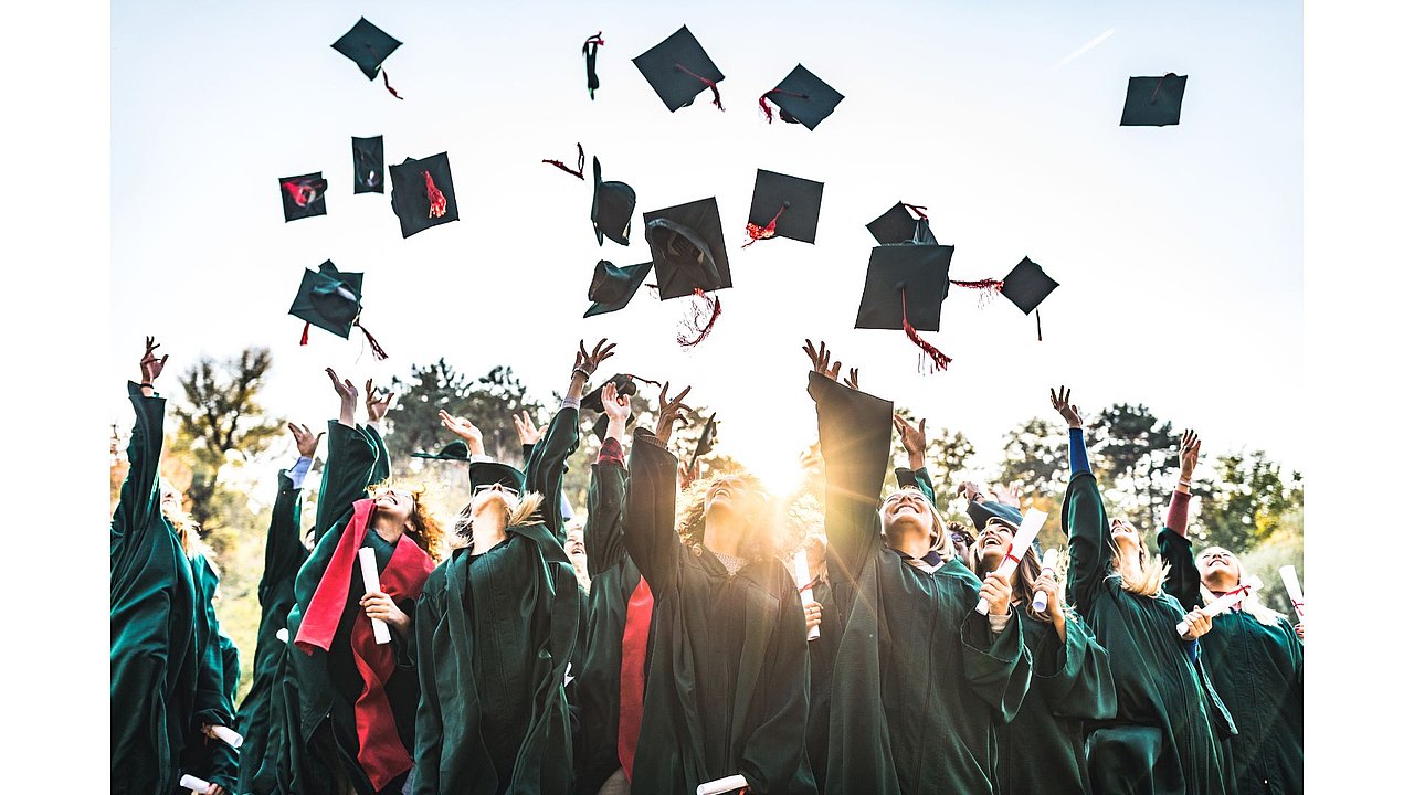 Gruppe von Absolventinnen und Absolventen in Talaren wirft bei einer Abschlussfeier ihre Hüte jubelnd in die Luft, während sie ihre Zeugnisse in den Händen halten.