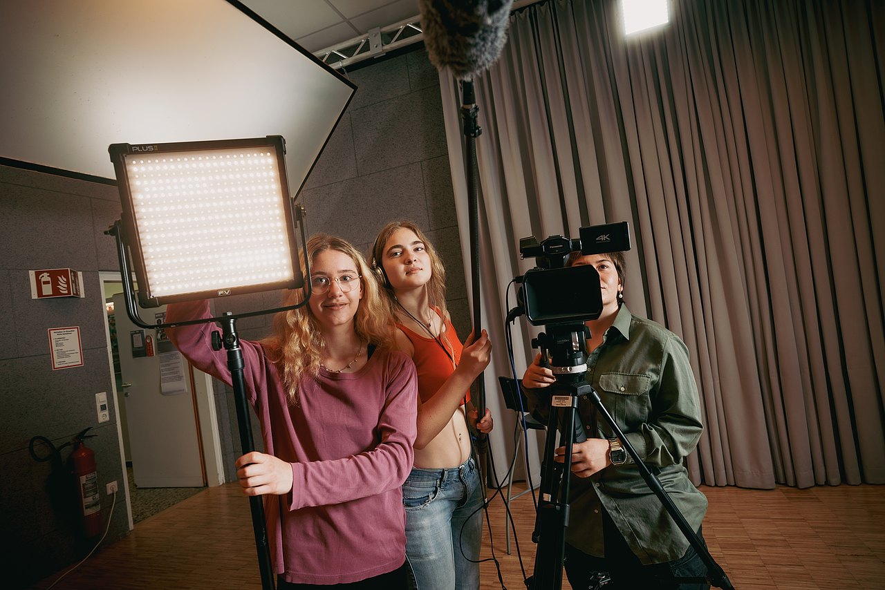 Eine Jugendliche bedient eine Panasonic-Videokamera in einem Studio, während eine andere Jugendliche eine LED-Flächenleuchte ausrichtet und eine dritte Jugendliche ein Mikrofon hält.