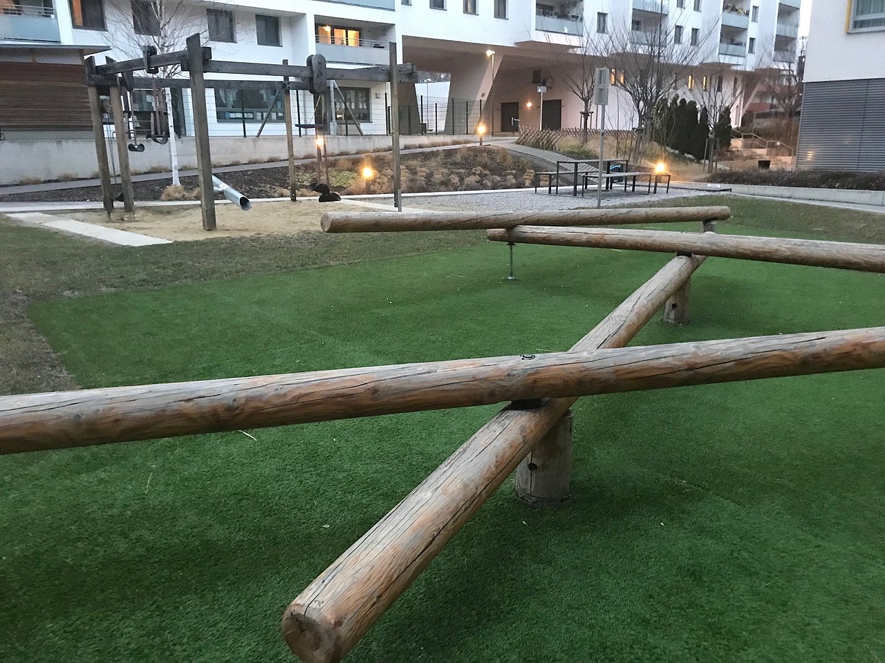 entrindete Baumstämme zum Balancieren übereinandergelegt, dahinter Schaukeln und Sandspielbereich 