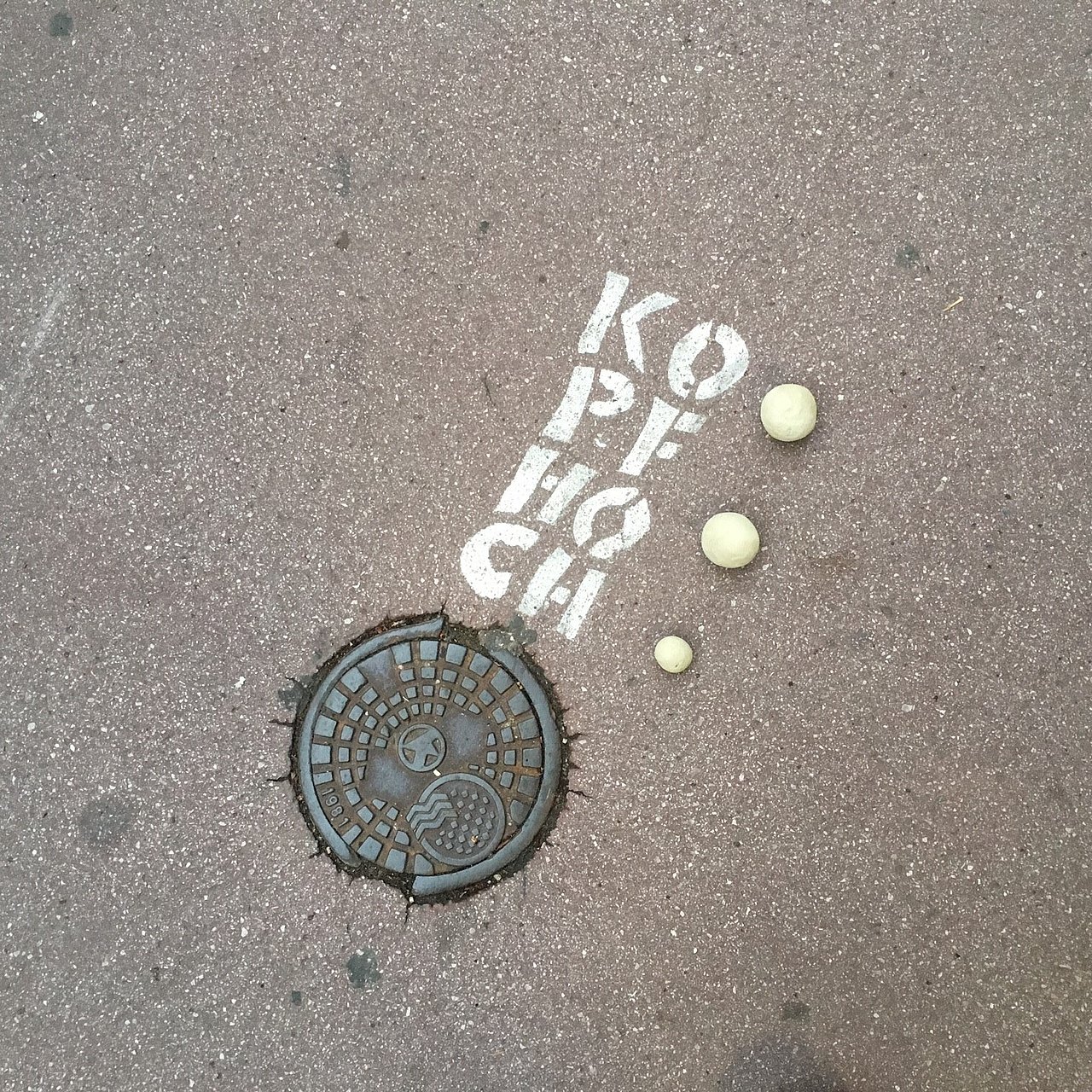 drei Lehmkugeln liegen auf Asphaltboden, daneben hat jemad gesprüht KO-PF-HO-CH