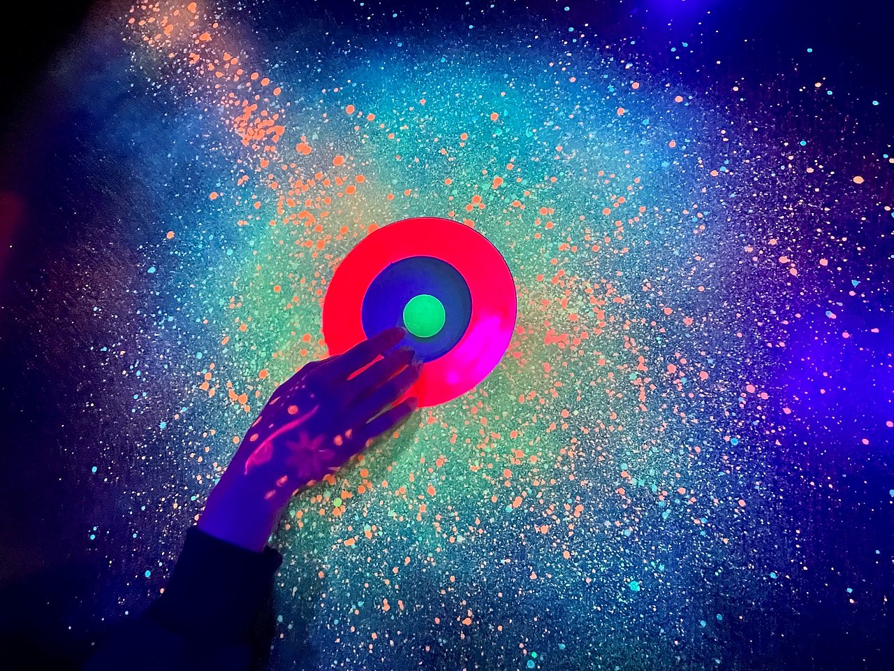 Hand mit Neonfarben bemalt, die gerade einen Minigolfball aus einem Loch holt, Umgebung schaut wie ein Sternenhimmel aus