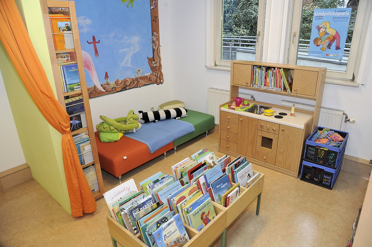 Ein Kinderbereich mit Bücherwand, einer Sitzbank mit Plüschtieren, darüber Wandbemalung. In der Mitte ist eine Holzküche, die auch mit Büchern gefüllt ist, daneben und davor weitere Bücherkisten mit Kinderbüchern.