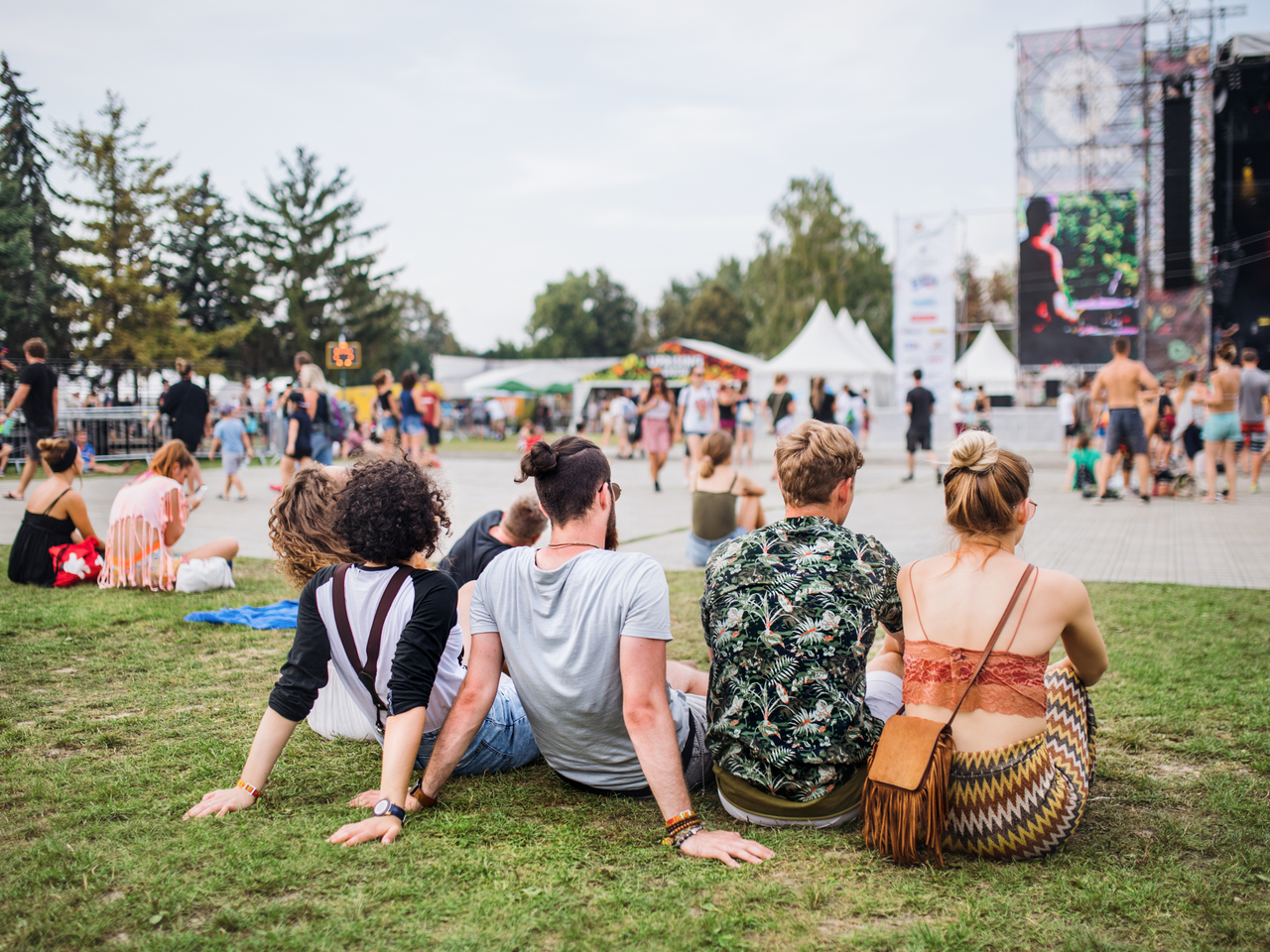 Das Bild zeigt eine Gruppe von vier jungen Erwachsenen von hinten, die entspannt auf einer Wiese sitzen und ein Open-Air-Event oder Musikfestival genießen.