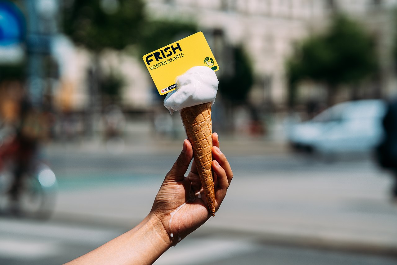 FRISH Vorteilskarte und Eiscreme: Eine Hand hält ein Eis in einer Waffeltüte, an dem eine gelbe FRISH-Vorteilskarte befestigt ist. Im unscharfen Hintergrund ist eine belebte Straßenszene mit Fußgängern, Autos und Gebäuden zu sehen.