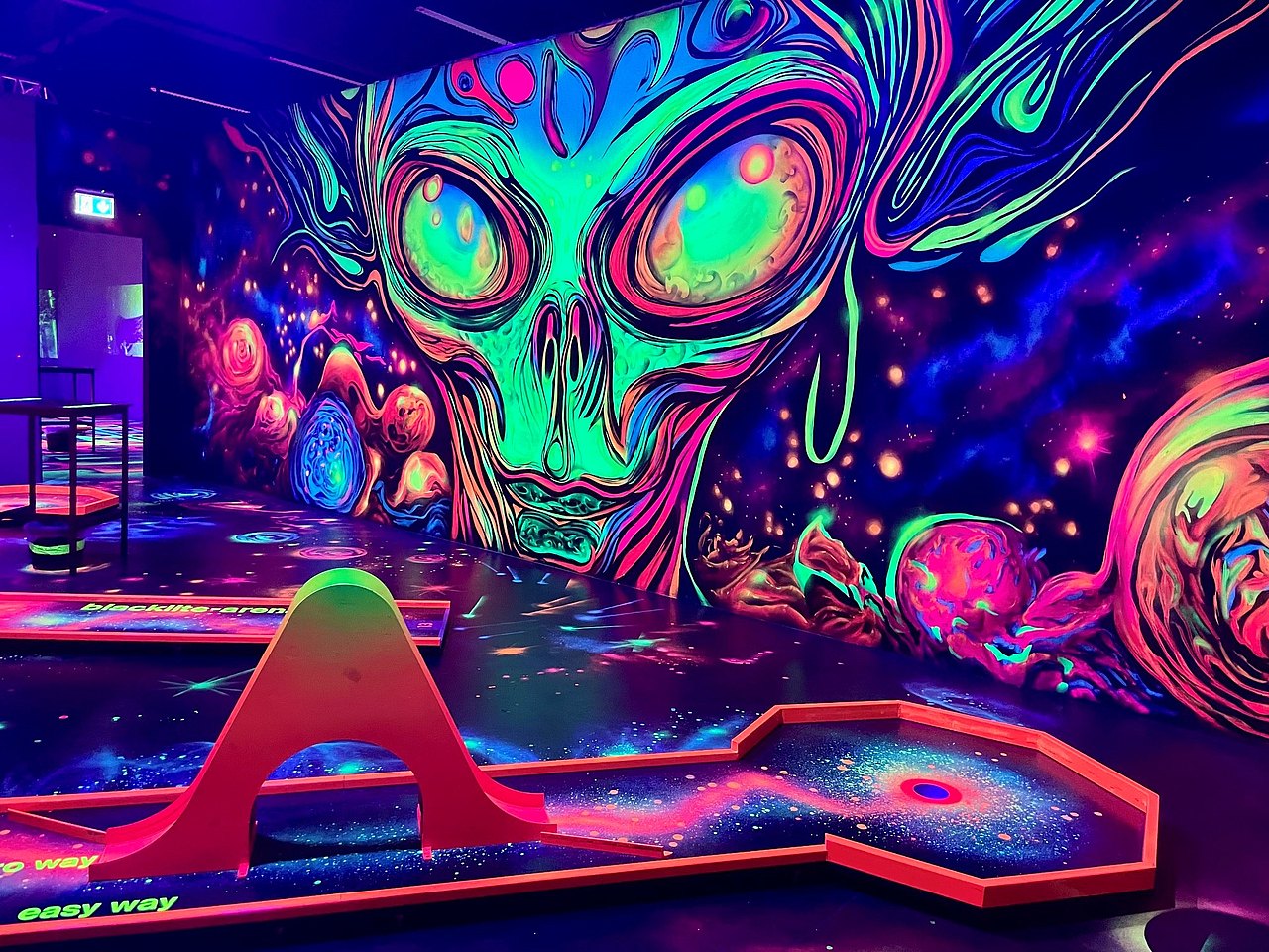 Neonfarbene Minigolfbahn im Hintergrund ein Gesicht, das dem Bild "Der Schrei" von Munch nachempfunden sein könnte