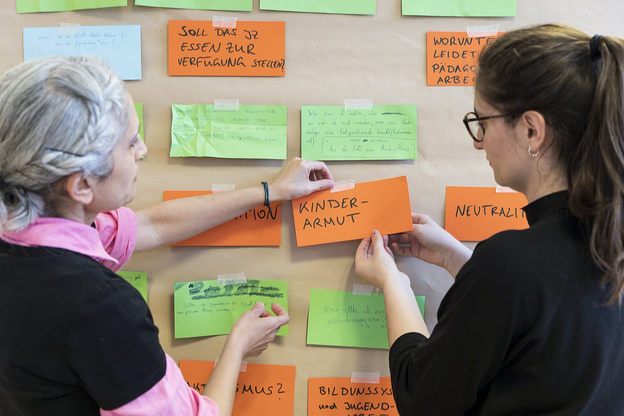 Zwei Frauen befestigen beschriftete Karten auf einer Pinnwand.
