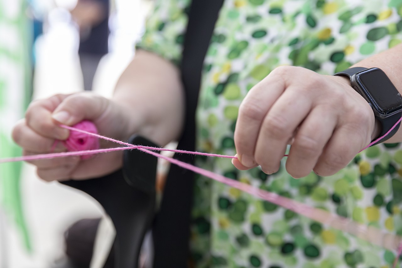 Die rechte und linke Hand einer weißen Person ziehen an zwei Enden von einem rosa Faden und bilden damit einen Konten.