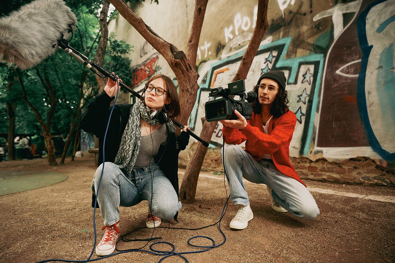 Zwei Jugendliche knien in einem Park am Boden, einer der Jugendlichen hält eine Videokamera, während die andere Jugendliche ein Mikrofon hält.