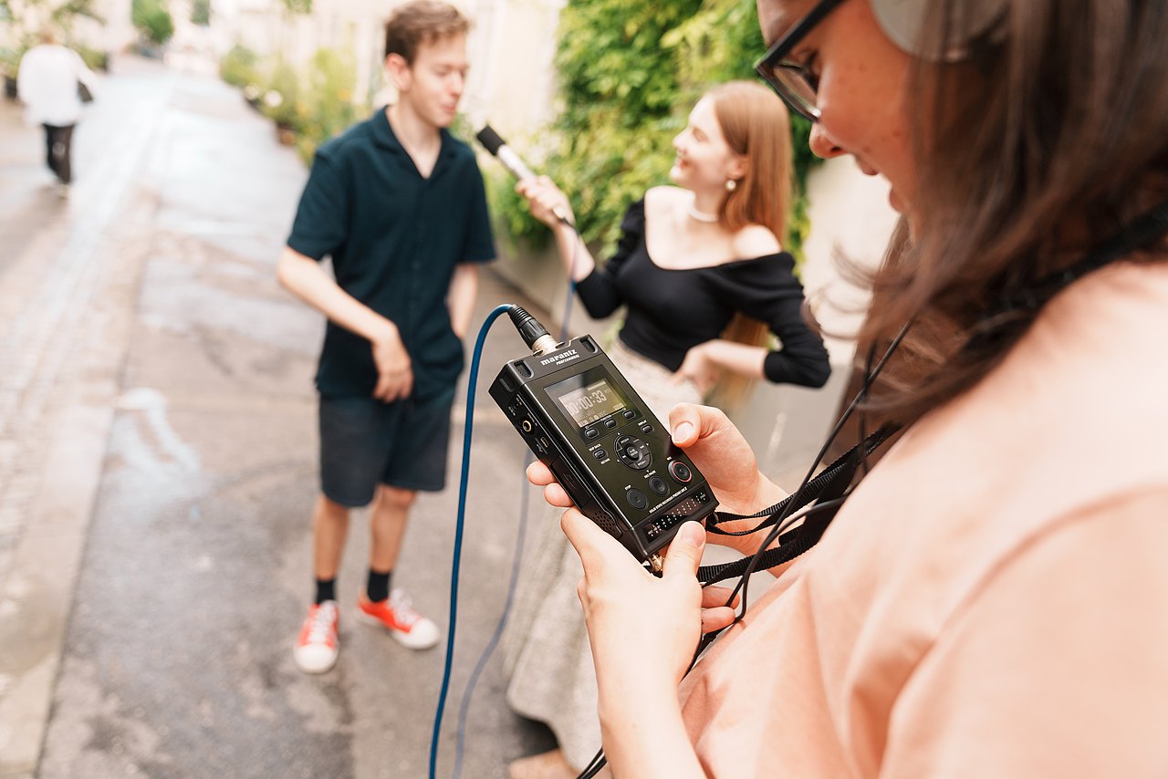 Ein Jugendlicher hält ein tragbares Audiorekorder-Gerät, während zwei andere Jugendliche ein Interview mit einem Mikrofon aufnehmen.