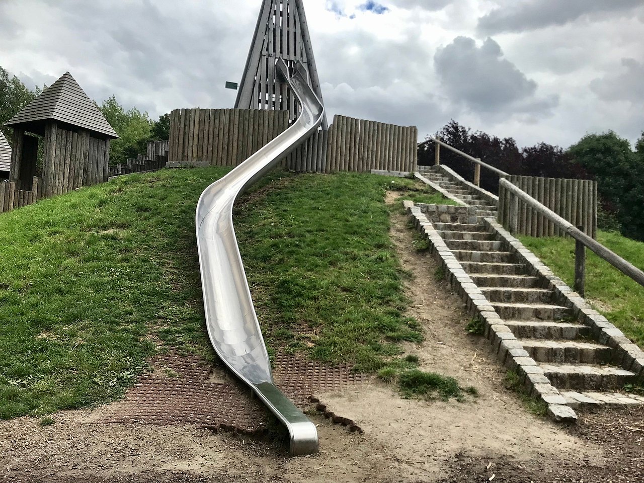 Alfred-Böhm-Park Holzgebäude auf einem grünen Hügel, wo rechts Steinstiegen mit Holzgeländer rauf führen. Aus dem Gebäude führt eine Metallrutsche runter.