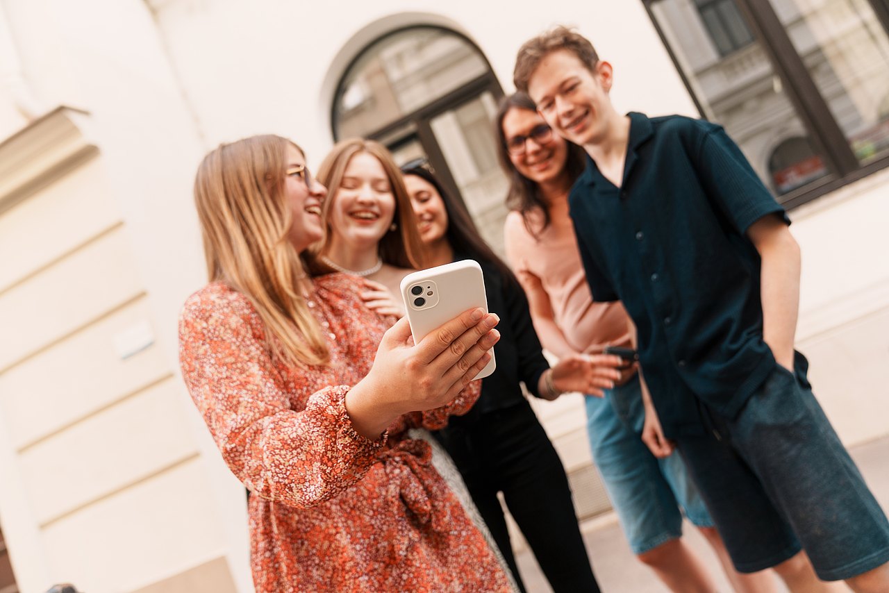 Eine Gruppe an lachenden Jugendlichen steht vor einem Haus in einem Kreis und schaut auf ein Handy..