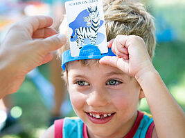 Zu sehen ist das Gesicht eines Kindes in Nahaufnahme. Das Foto entstand auf einer Spieleveranstaltung im Park. Das Kind spielt das Spiel Hedbanz und trägt ein Stirnband, in das eine Karte mit der Abbildung eines Zebras gesteckt ist.