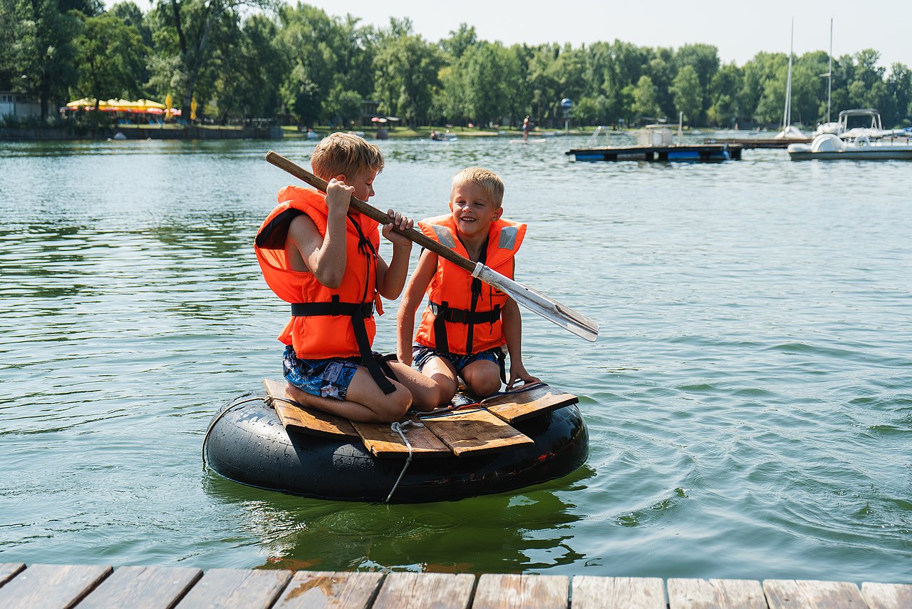 2 Kinder mit Schwimmwesten auf einem Floss im Wasser