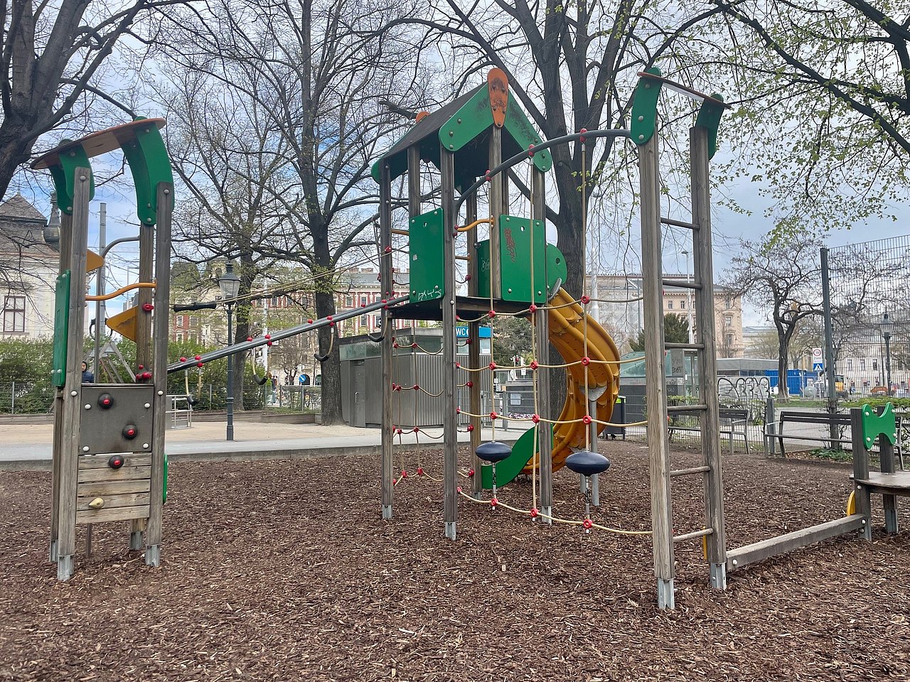 großes Spielgerät mit vielen unterschiedlichen Klettermöglichkeiten und gebogener Rutsche, im Hintergrund Toilettenanlage und Karussel