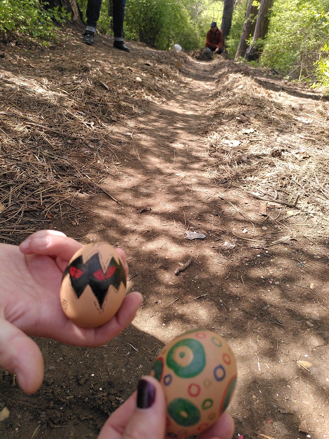 Erdhügel von oben. Zwei Hände halten zwei bemalte Eier in die Kamera.