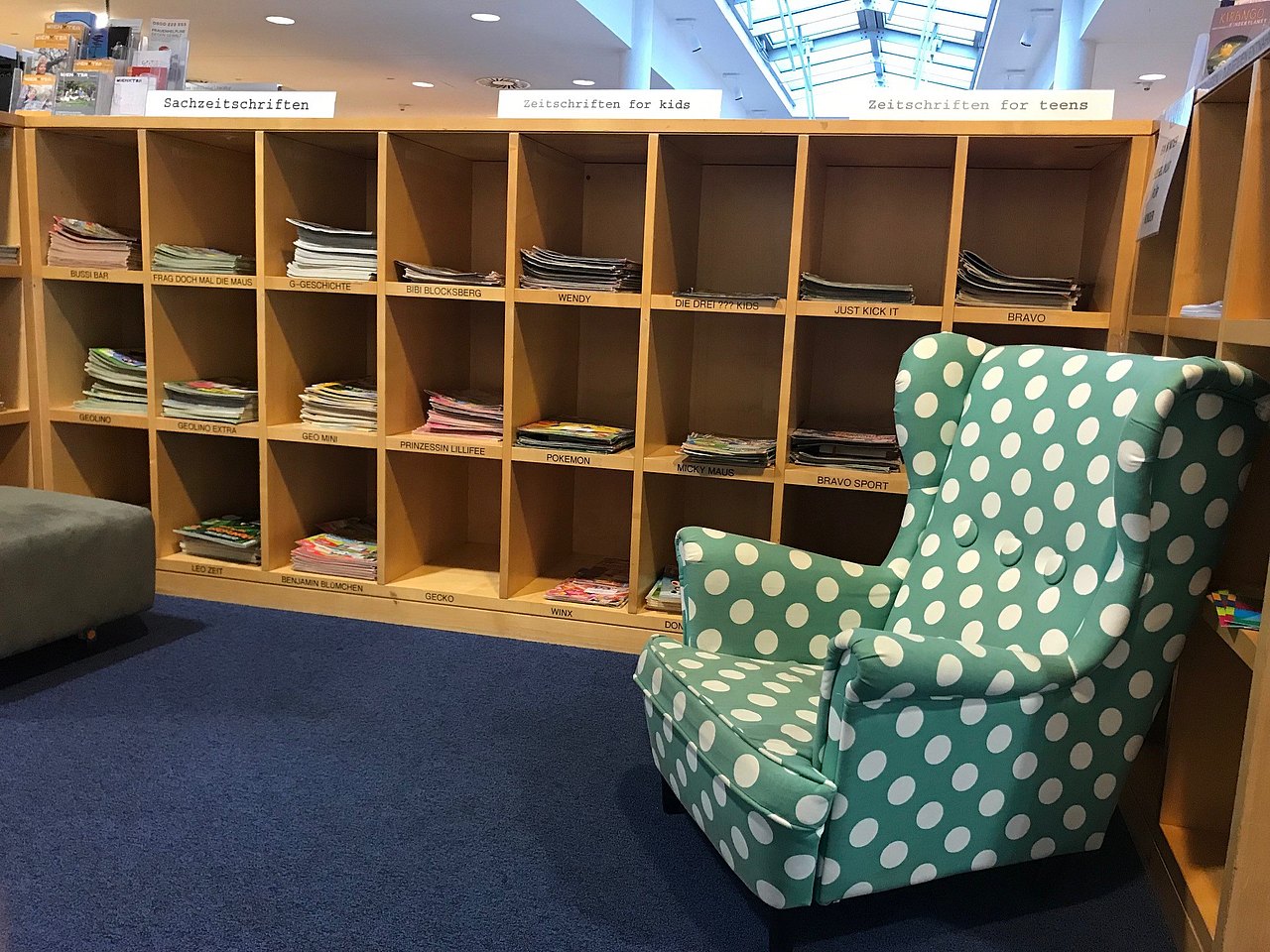 Ein Grüner Ohrensessel mit weißen Punkten vor einem Zeitschriftenregal.