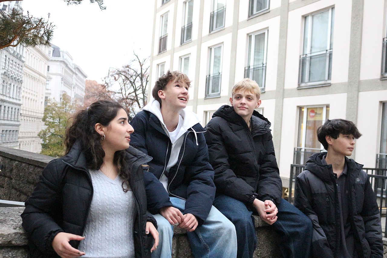  Vier junge Erwachsene, die draußen auf einer Straße vor hellen Gebäuden sitzen und stehen.