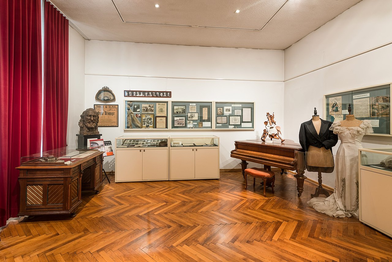 Ausstellungsraum des Bezirksmuseums Josefstadt mit Bildern an der Wand, Schaukästen, einer Büste, einem Klavier sowie einem Brautkleid und Jackett.