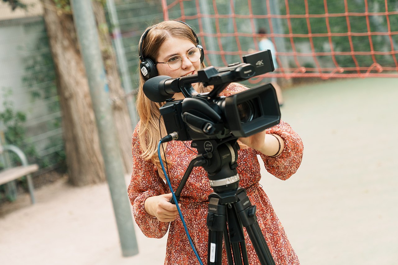 Eine Jugendliche mit Kopfhörern filmt im Freien mit einer Panasonic-Videokamera auf einem Stativ.