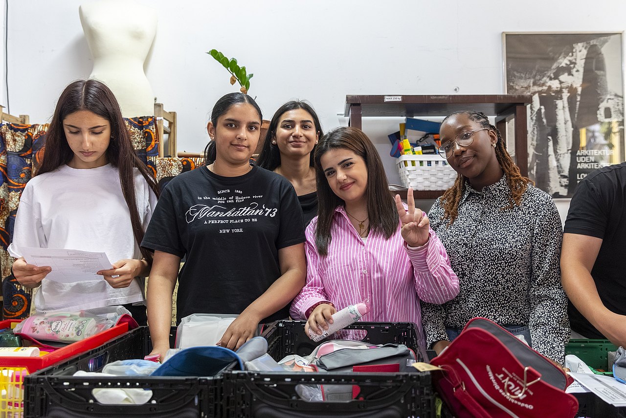 Sechs lächelnde Personen stehen hinter verschiedenen Kleidungsstücken und Accessoires im Vordergrund und Hintergrund.