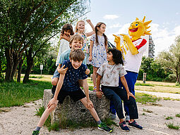 Ferienspiel-Maskottchen Holli lacht gemeinsam mit 6 Kindern.