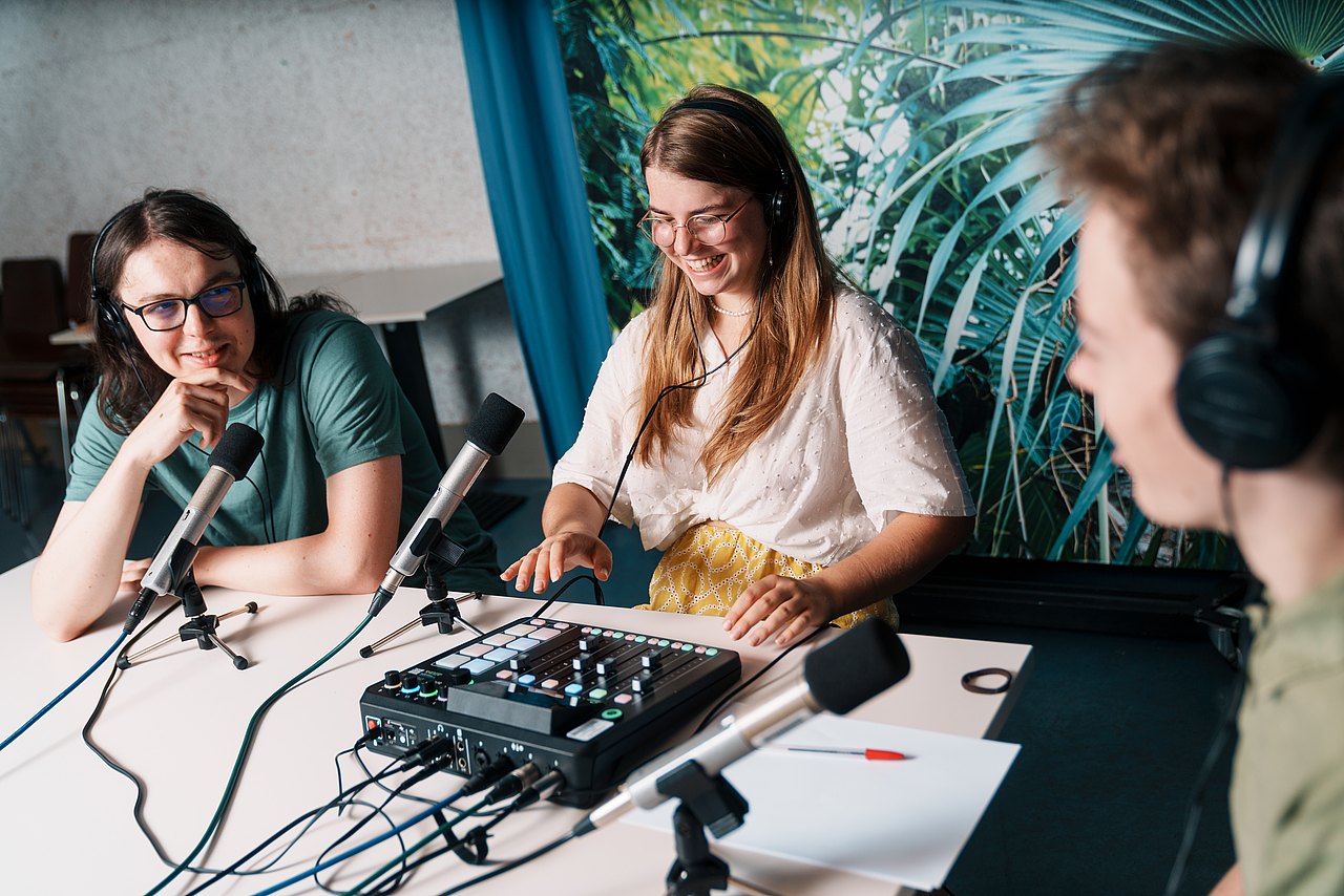 Drei Jugendliche sitzen an einem Tisch mit Mikrofonen und einem Audio-Mischpult und nehmen gemeinsam einen Podcast auf..