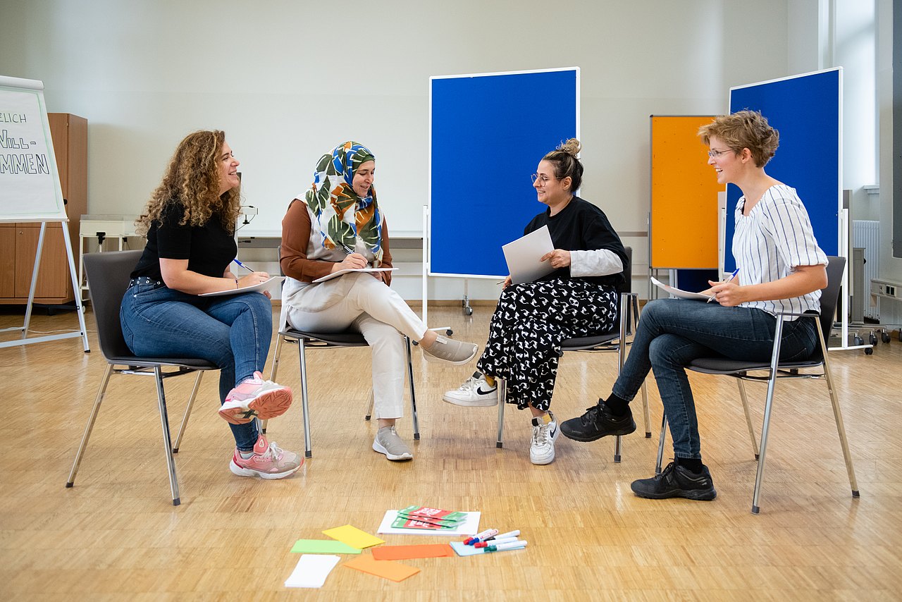 Vier Frauen sitzen auf Sesseln im Kreis im Seminarraum und sprechen miteinander. Am Boden liegen Moderationskärtchen und im Hintergrund sind verschiedenfarbige Pinnwände aufgestellt
