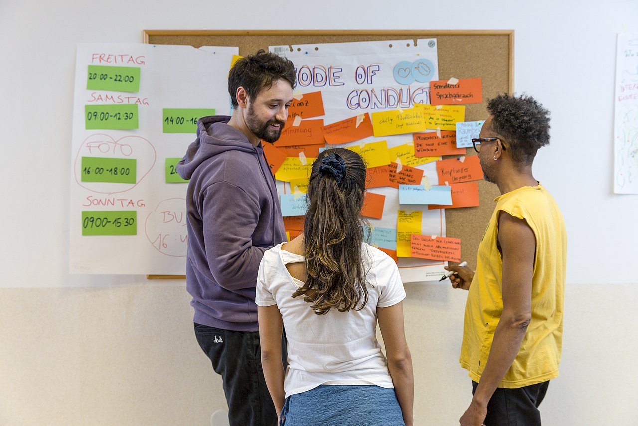 Drei Personen unterhalten sich vor einer Pinnwand, auf der Post-its hängen