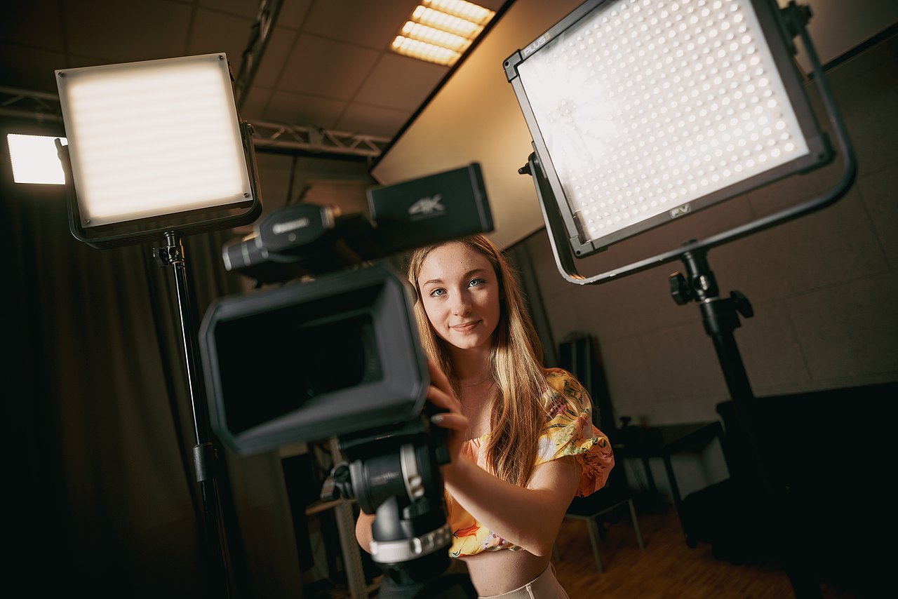 Eine Jugendliche steht hinter einer Kamera auf einem Stativ und zwei Videoleuchten in einem Raum.