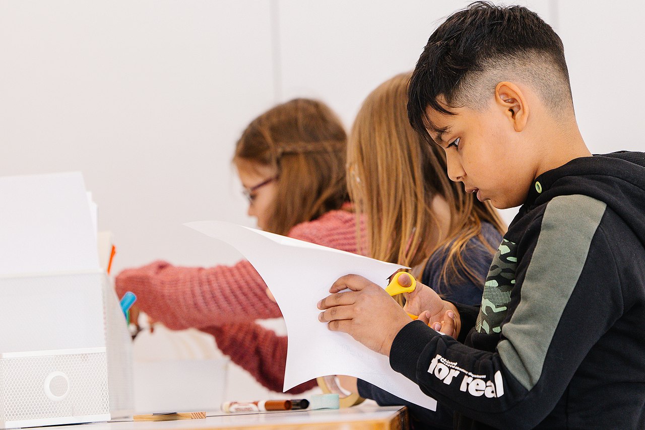 Kind schneidet Blatt Papier, dahinter zwei Kinder am kreativ arbeiten