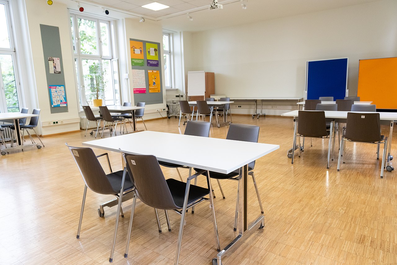 Miete dich bei uns ein! Ein heller Seminarraum mit großen Fenstern und mehreren Tischen, Stühlen, Pinnwänden und einem Flipchart.