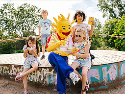 Ferienspiel-Maskottchen Holli blättert gemeinsam mit Kindern im Programm vom Ferienspiel. 