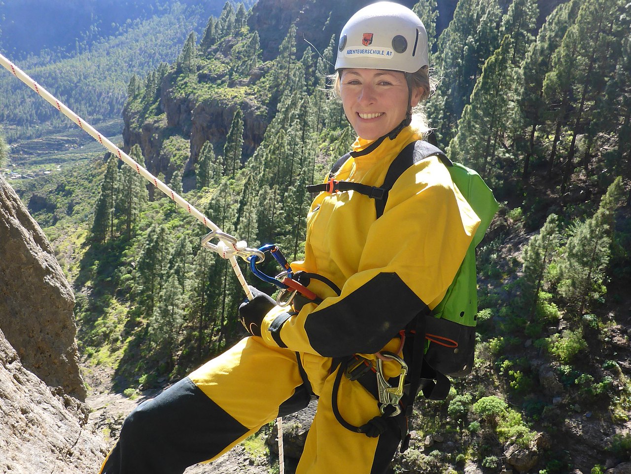 Eine lächelnde Frau beim Abseilen an einer felsigen Steilwand. Sie trägt einen leuchtend gelben Canyoning-Anzug mit schwarzen Einsätzen, einen weißen Schutzhelm, Kletterhandschuhe und einen grünen Rucksack. Sie ist über ein Kletterseil mit Karabinern gesichert, das sie mit einer Hand hält. Im Hintergrund erstreckt sich eine weite, sonnige Berglandschaft mit vielen grünen Nadelbäumen.