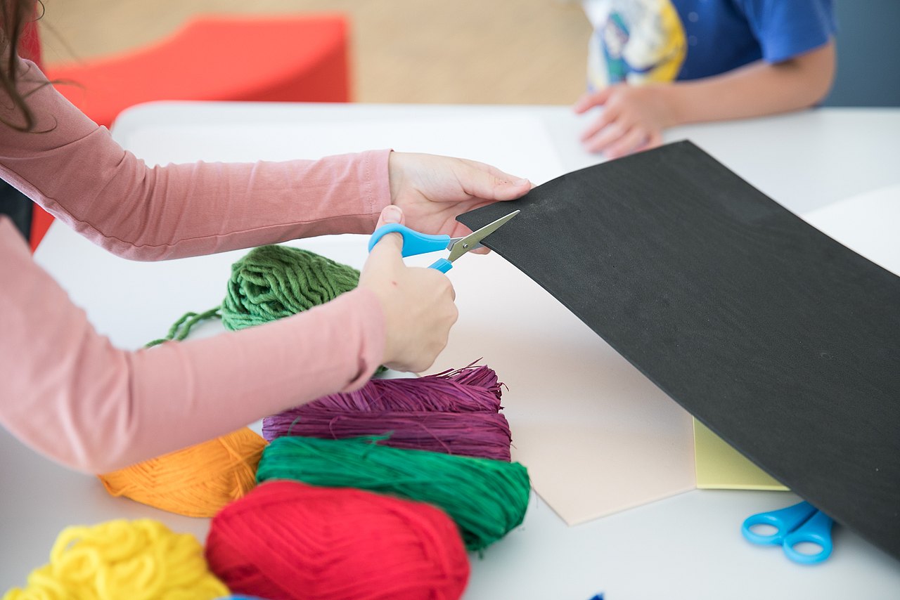 Kind schneidet mit Schere Blatt Papier, Wollknäuel liegen auf dem Tisch