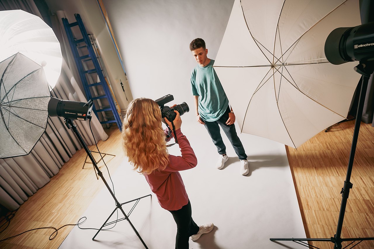 Eine Jugendliche fotografiert einen Jugendliche vor weißem Hintergrund in einem Studio.