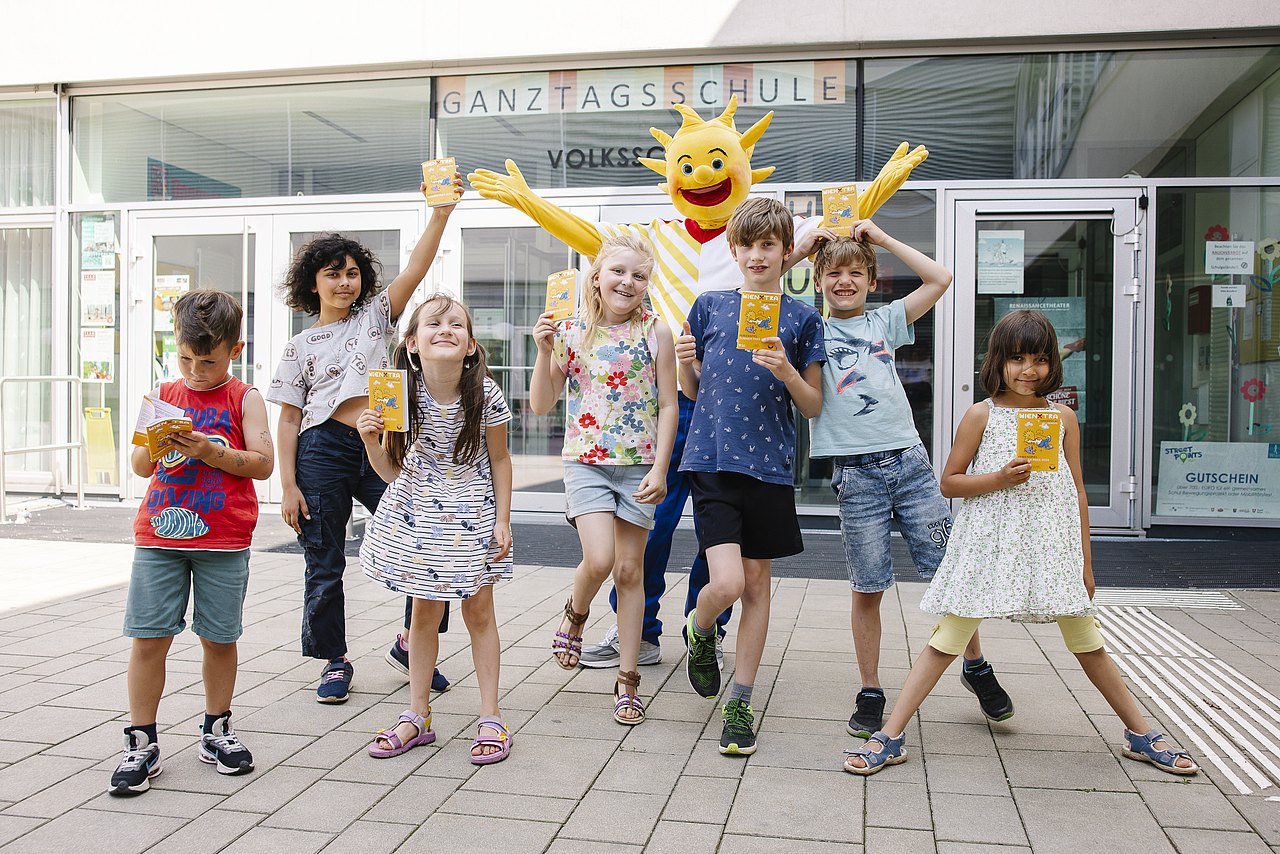 fröhliche Kinder und Ferienspiel-Maskottchen Holli vor einer Schule