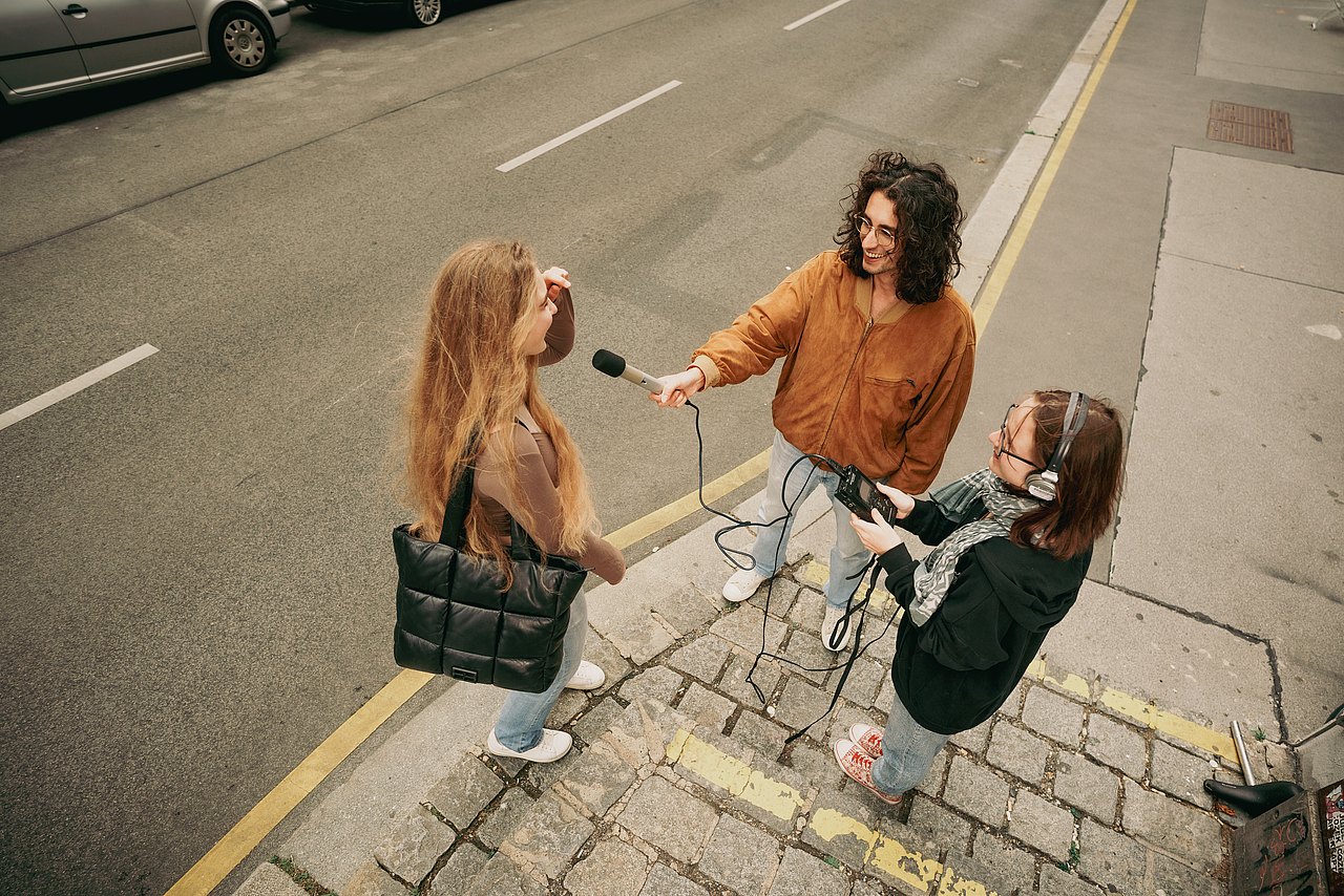 Drei Jugendliche stehen am Gehweg, einer hält ein Mikrofon und interviewt die andere Person.