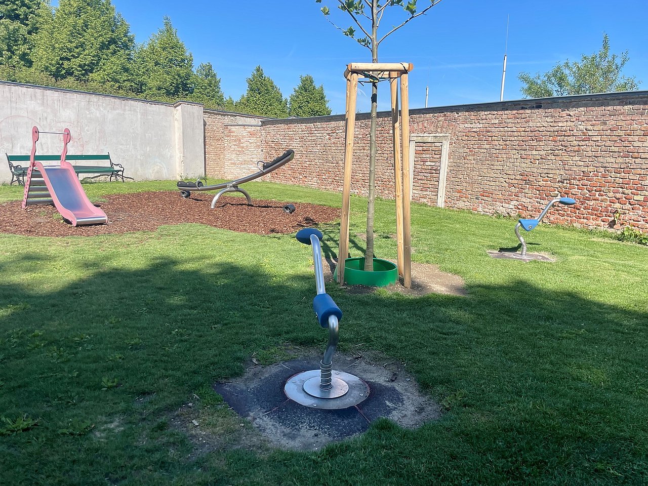 Spielgeräte am Spielplatz im Belvedere Garten Kleinkidnerrutsche, Wippe und eine Bank im Hintergrund die Backsteinauer