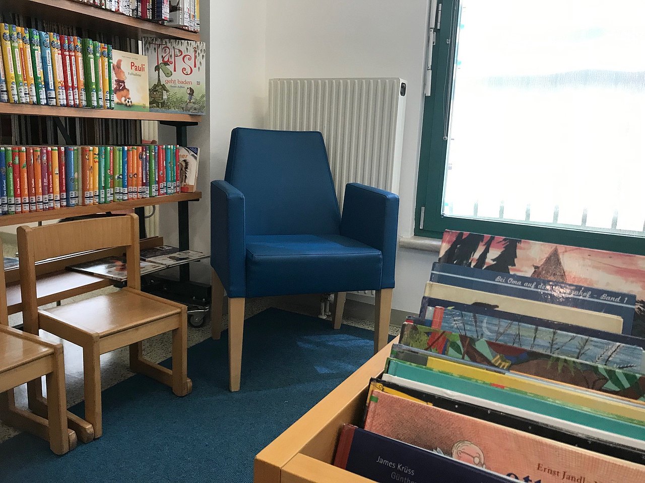 Ein blauer Sessel im Eck, daneben zwei Holzsessel, dahinter eine Kinderbücherwand und davor eine Kiste mit Kinderbüchern.