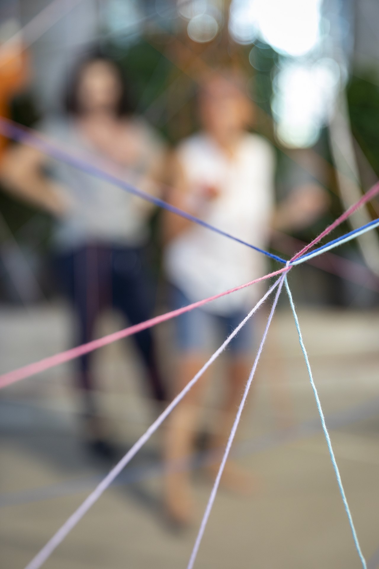 Im Hintergrund sieht man verschwommen zwei Personen, im Vordergrund ist ein Spinnennetz aus bunten Fäden zu sehen.