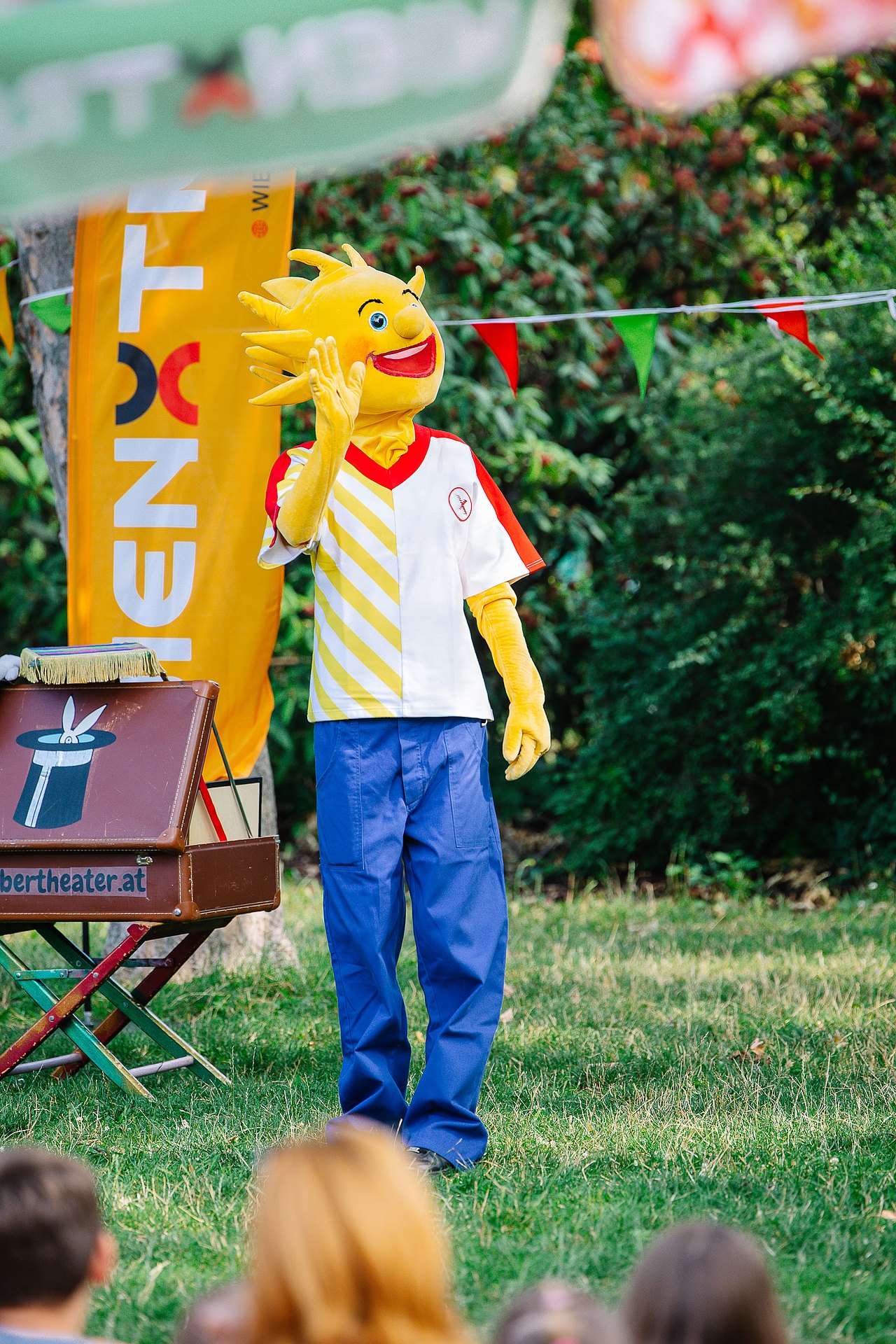 Das Kinderaktiv-Maskottchen Holli steht im Park neben einem Zauberkoffer vor einer Wienxtra-Flagge und bunten Wimpeln und winkt