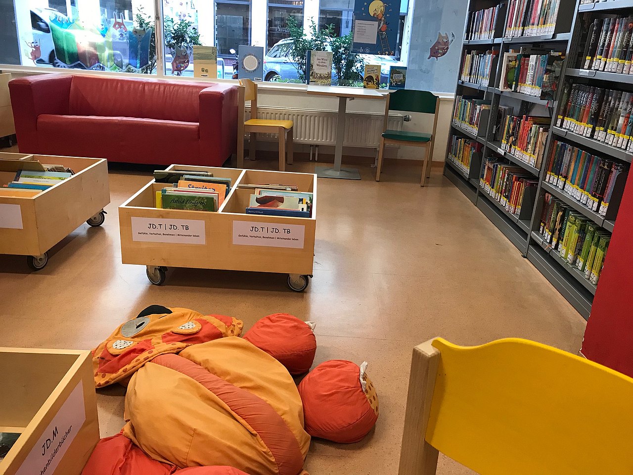 Rechts eine Bücherwand, zwei Holzsessel mit Tisch und eine rote Couch. In der Mitte vom Raum sind Bücherkisten und ein Sitzsack.