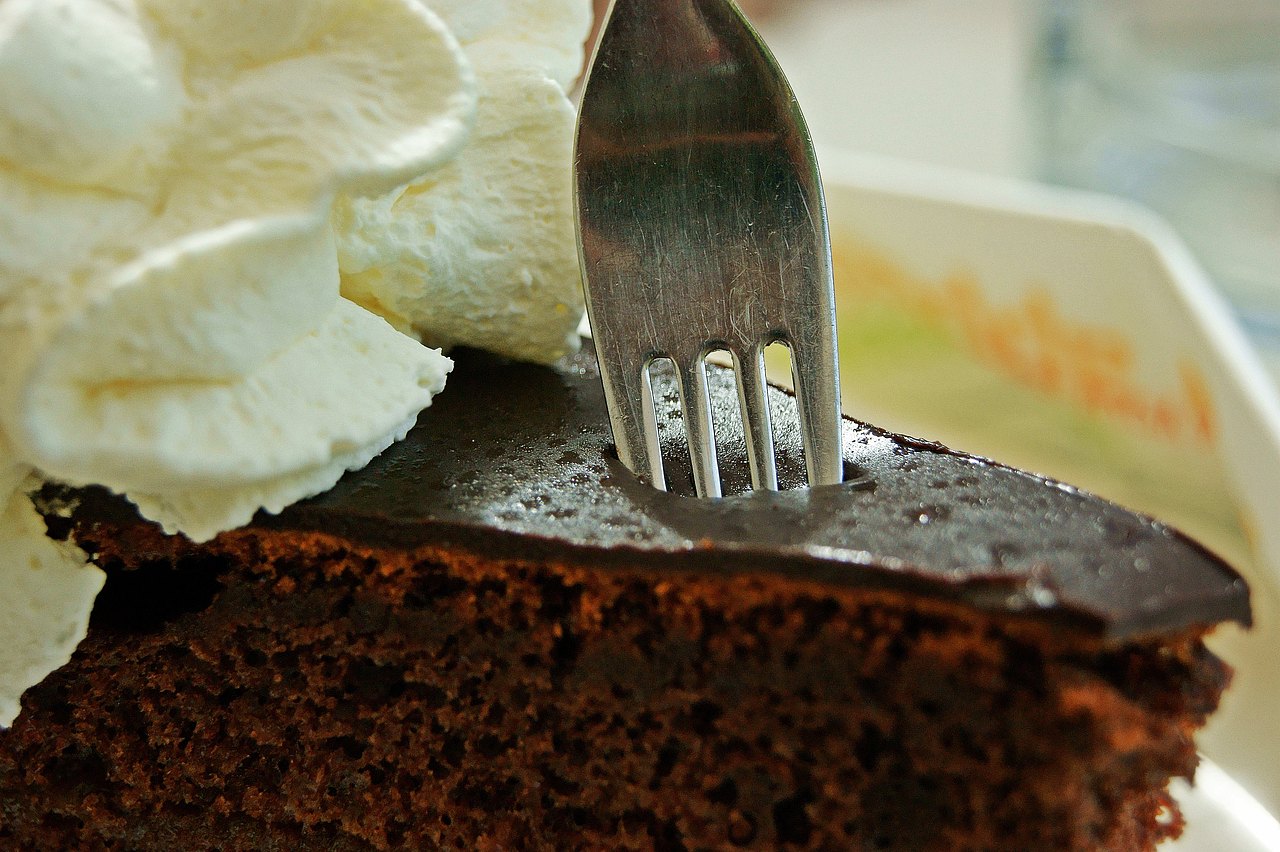 Eine Gabel sticht in eine Sachertorte. Die Torte ist dekoriert mit Schlagobers. 