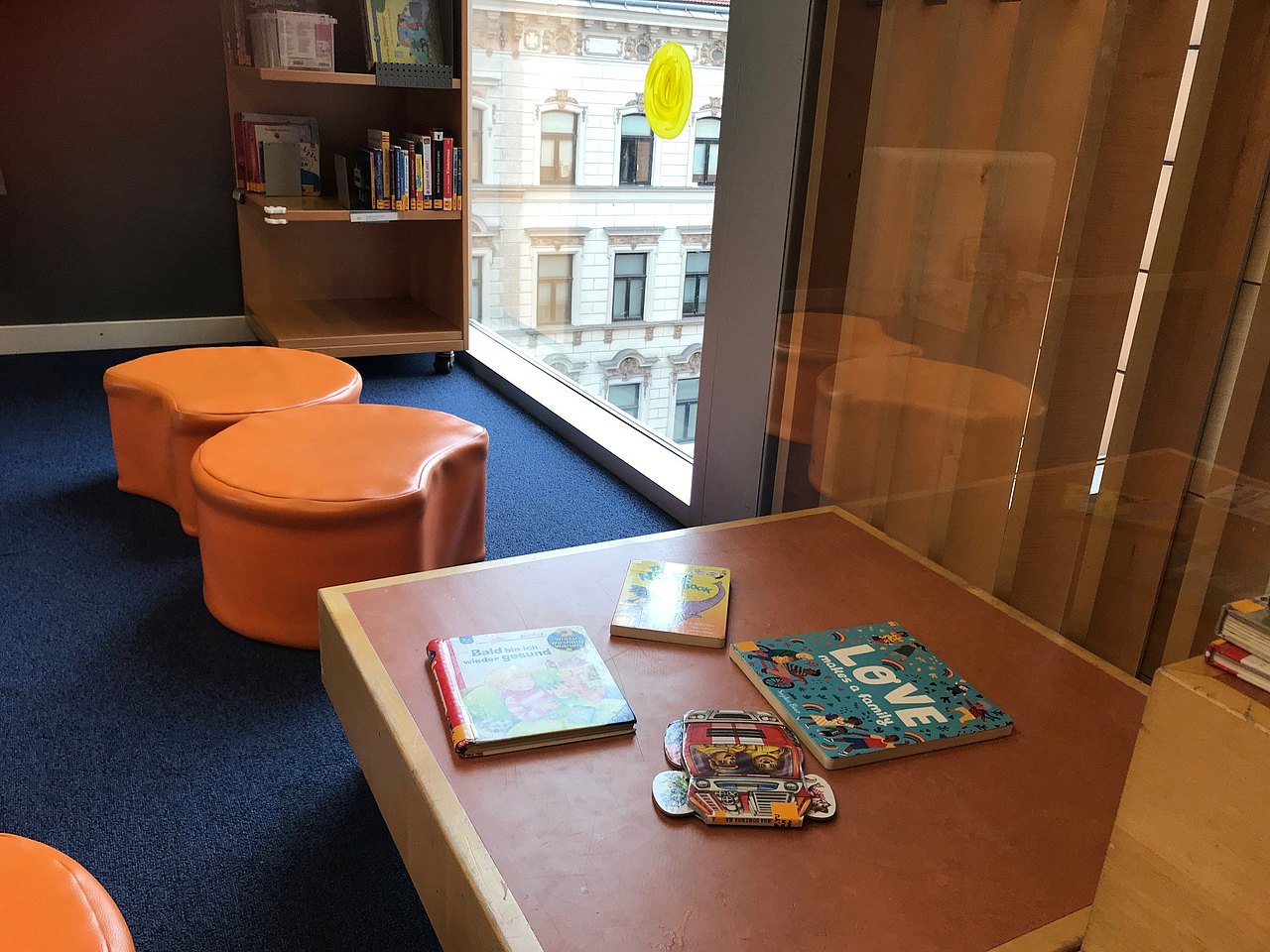 Orangene Sitzhocker auf blauem Teppichboden. Ein kleines Bücherregal und vier Kinderbücher auf einem Holztisch.
