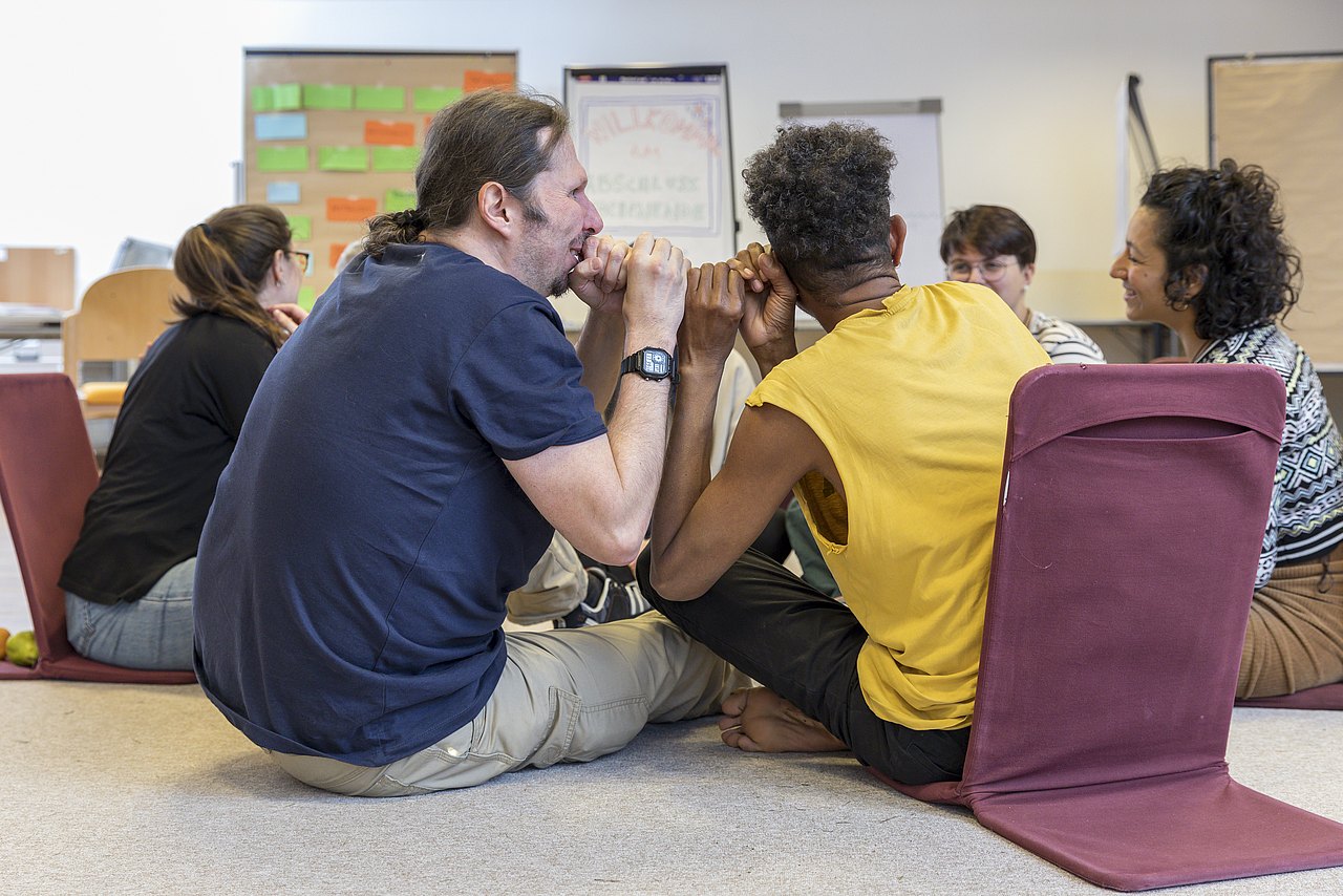 Mehrere Personen sitzen in einem lockeren Kreis auf dem Boden. Sie nutzen bodennahe Sitzstühle mit Rückenlehnen. Im Hintergrund sind Flipcharts und Pinnwände mit Notizen zu sehen, was auf eine Brainstorming-Session oder einen Workshop hindeutet. Die Teilnehmenden sind in ein Gespräch vertieft.