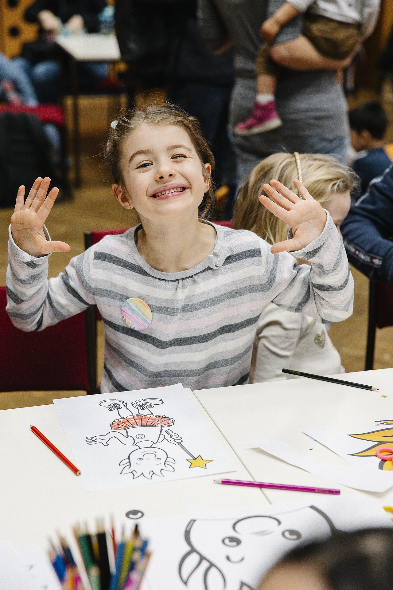 breit lachendes Mädchen mit teils angemalten Malvorlagen an einem Tisch, beide Hände in die Luft gestreckt