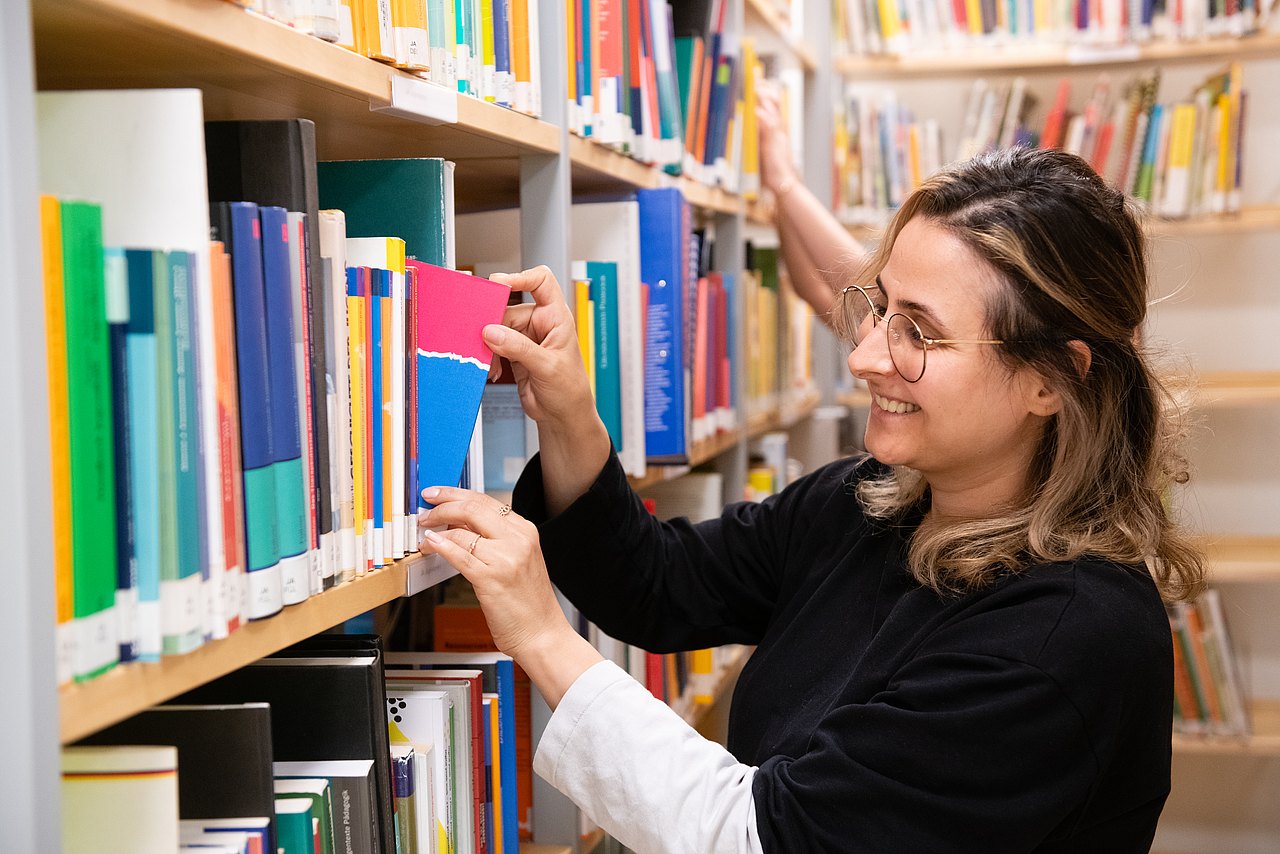 Eine Frau steht vor einem Bücherregal in der Bibliothek und greift mit ihrer rechten Hand nach einem Buch.