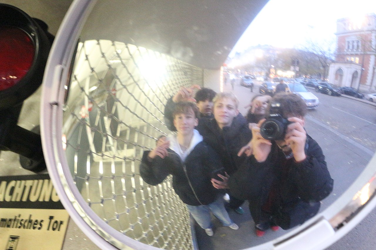 Ein Jugendlicher fotografiert einen runden Straßenspiegel, in der Reflexion sieht man ihn und seine Freunde.