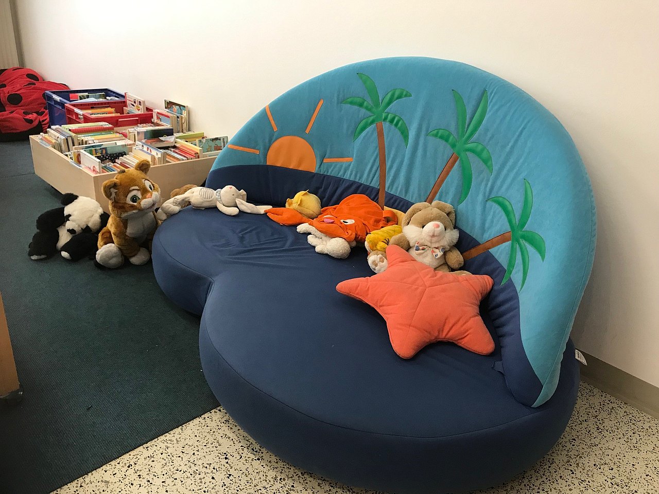 Eine Sitzbank in Kindergröße, mit Palmen- und Sonnenmotiv. Darauf und daneben sind verschiedene Plüschtiere platziert. Daneben sind Bücherkisten mit Kinderbüchern.